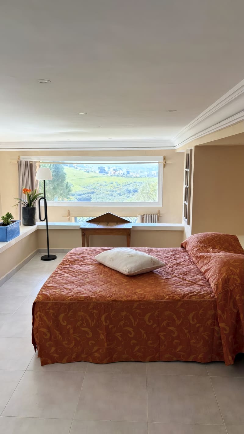 Chambre lumineuse avec lit au couvre-lit orangé, face à une large fenêtre donnant sur la colline verte.