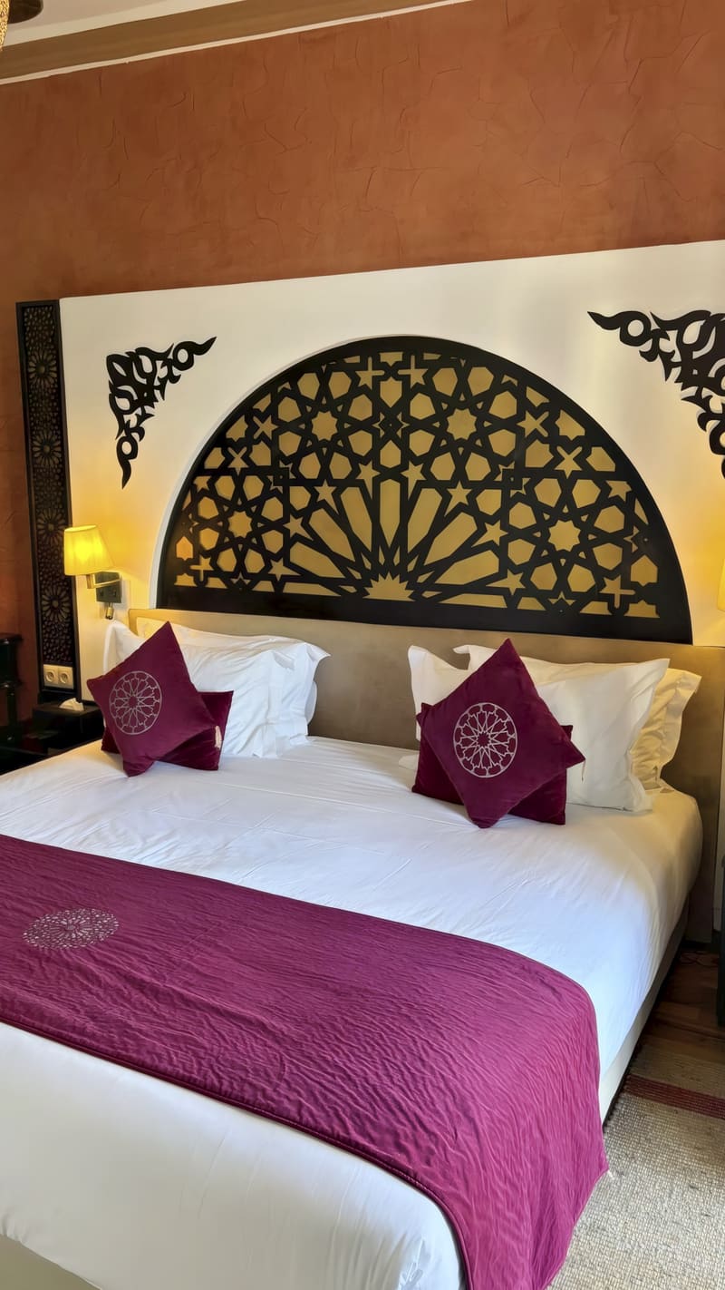Lit double aux draps blancs et coussins pourpres, sous une tête de lit en demi-cercle au motif géométrique ajouré, murs couleur terre cuite.