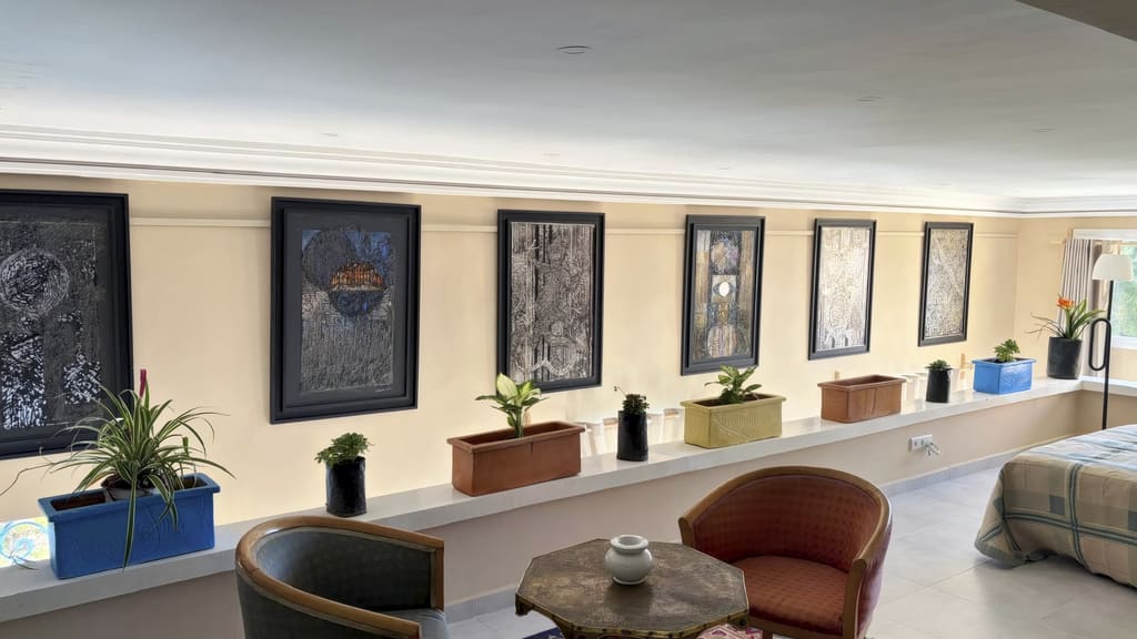 Grande pièce aux murs clairs : une rangée de toiles encadrées, des jardinières sur le rebord, deux fauteuils autour d'une table octogonale et un lit sur la droite.
