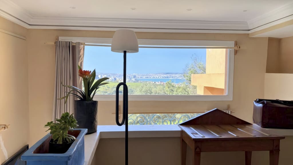 Grande fenêtre horizontale ouvrant sur la baie de Tanger, une plante fleurie et une lampe au premier plan.